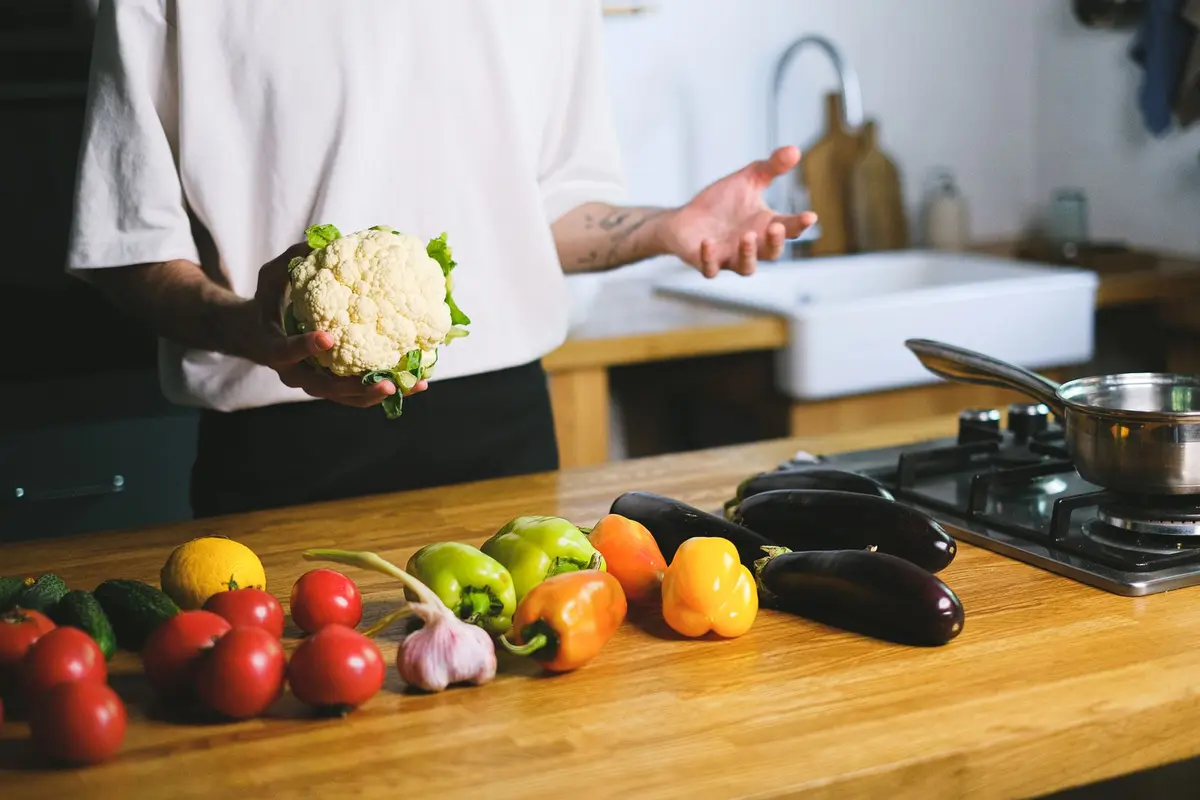 Hands holding a cauliflower over a kitchen counter with peppers, tomatoes and eggplants.